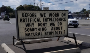 Image of a strip mall sign that reads: Before we work on artificial intelligence, why don't we do something about natural stupidity? This is probably the most cyberpunk thing I've seen all day. 